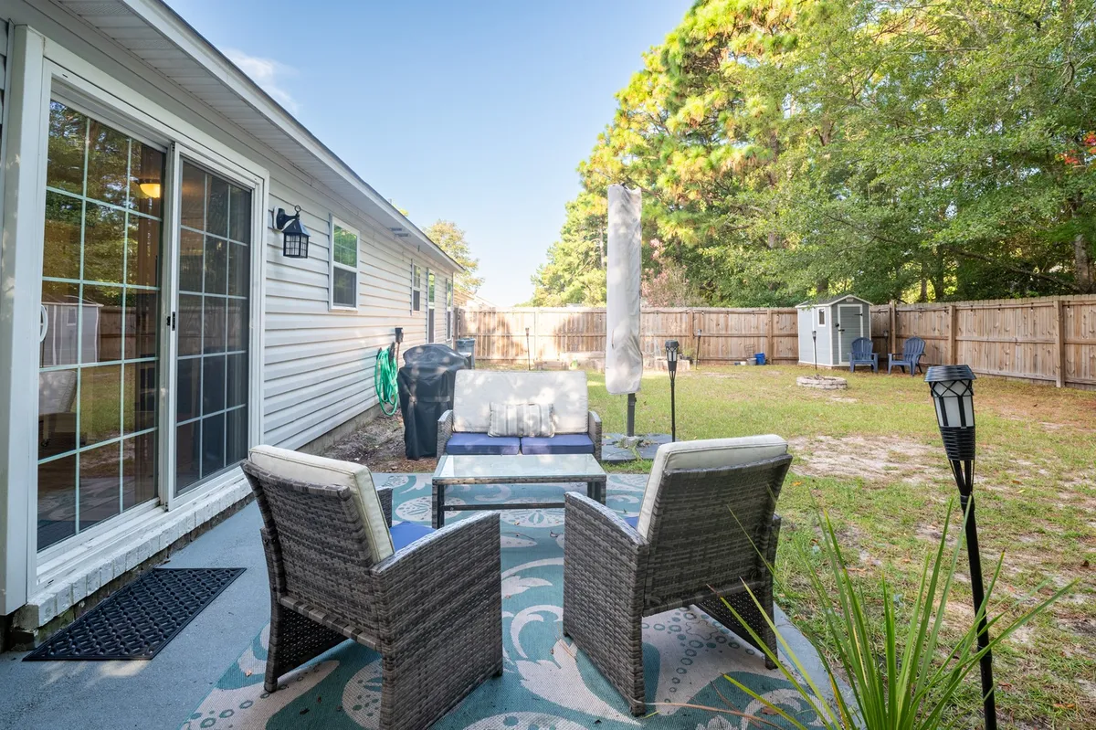 The image captures an outdoor seating area featuring a glass table, chairs, lush backyard greenery, and a visible fence. The space feels comfortable and welcoming, suggesting meals or relaxation outdoors within a safe, contained area.