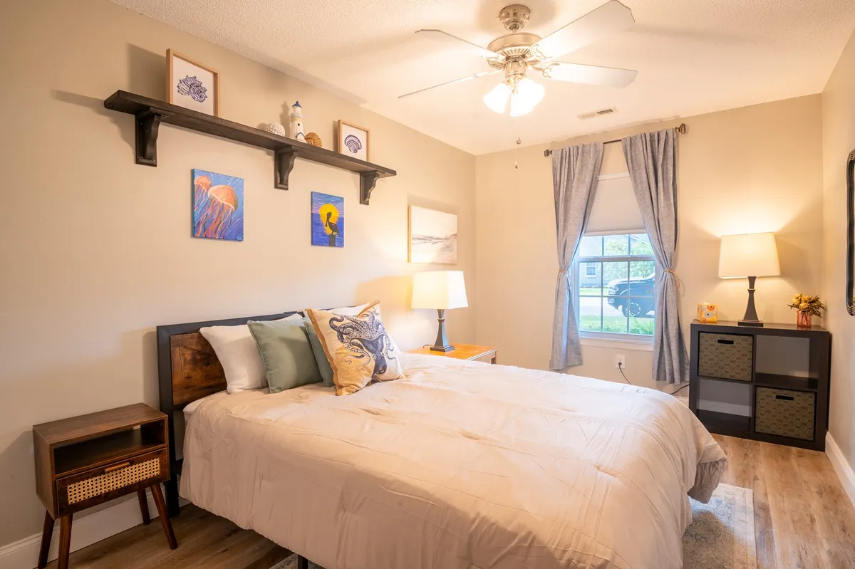 A sunlit bedroom featuring a bed with decorative pillows, wall art depicting marine life, and a window letting in natural light. The serene and cozy setting suggests a peaceful environment ideal for guests needing rest. The overall décor and soft ambiance make it a versatile space for friends or family.