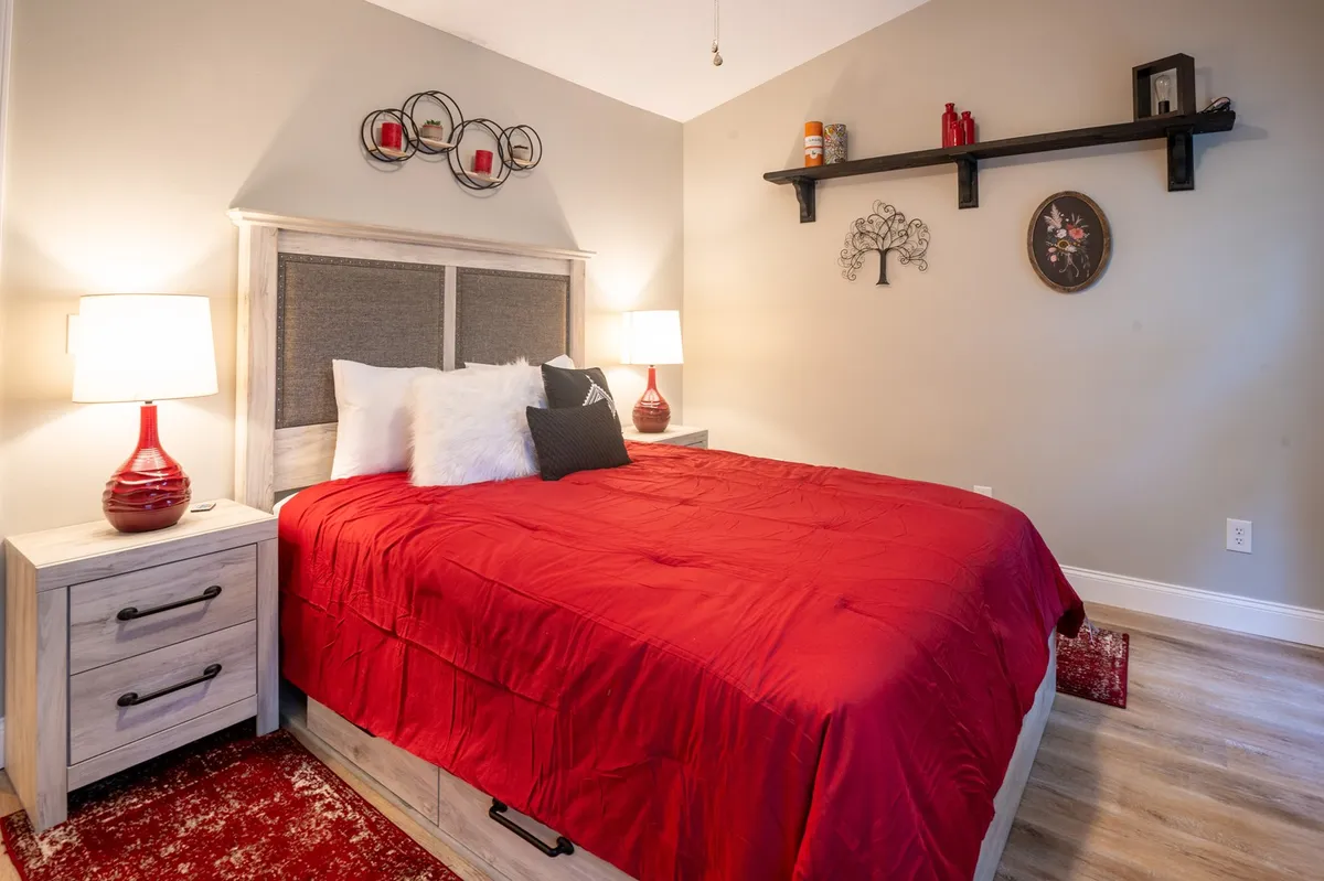 This image depicts a bedroom with a red bedspread, flanked by nightstands with lamps and a decorative wall shelf above. The room radiates an inviting atmosphere ideal for relaxation, reading, or sleep. The cozy setup, soft lighting, and thoughtful décor create a sense of tranquility and privacy, aligning well with the master suite description.