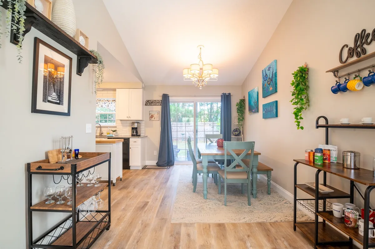 A warm, welcoming dining area with a table set for six, highlighted by a chandelier above, tasteful artwork on the walls, green plants, and a convenient coffee station. The ambiance is both functional and elegantly casual, suitable for shared meals and gatherings.