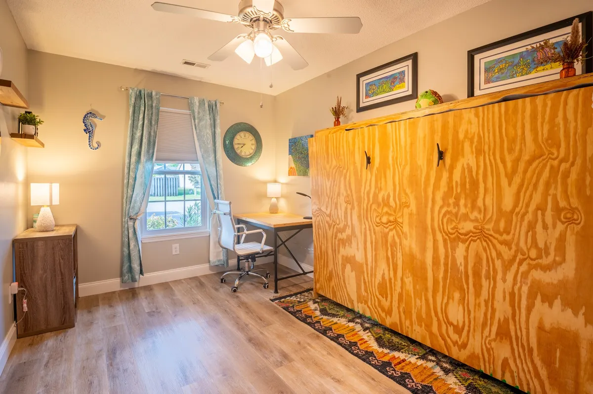 A cozy and productive workspace featuring a desk with a comfortable chair, wooden cabinet, window with curtains, decorative wall art, and a table lamp. The setup is well-lit, functional, and welcoming, supporting both focused work and moments of relaxation.