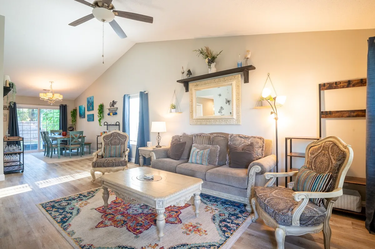 A spacious and comfortable living room featuring a soft, inviting sofa, a decorative coffee table, a colorful area rug, and a large mirror that reflects light and space. In the background, the dining area is visible, suggesting a seamless connection between lounging and dining spaces.