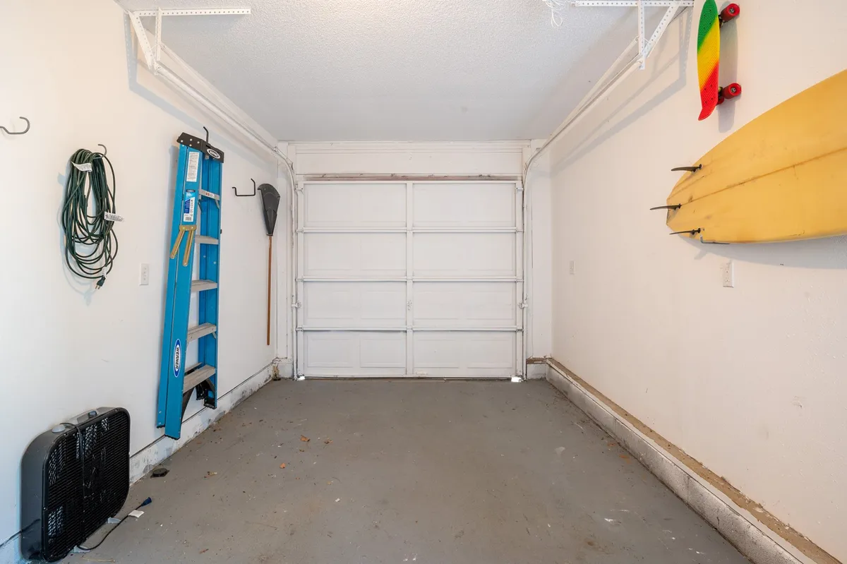 An interior view of the garage space, complete with organized wall-mounted hooks, a blue ladder, a yellow surfboard, and a green hose. The space is clean and functional, suggesting ample storage for personal items or recreational gear.