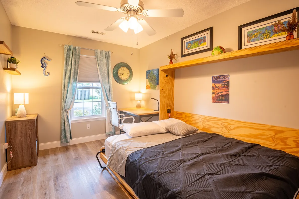 A multi-purpose room with a bed dressed in a black comforter, a desk and chair beside a window with curtains, and wall art adorning the space. This setup offers both a cozy sleeping area and a functional workspace, epitomizing the flexible use of the office/guest room with a Murphy bed.